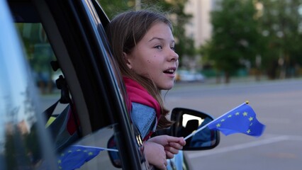 Close up shot of little pretty kid looking out car window with flag of European Union. Small girl holds EU flag on family car trip. Traveling in Europe, journey, slow motion concept