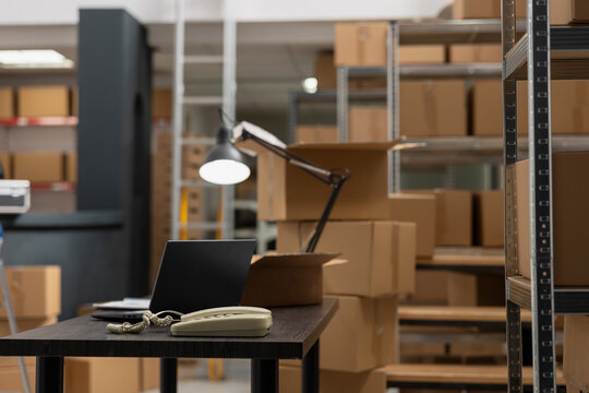 Storage room shipping area with laptop in empty fulfillment center, various taped boxes placed on shelves and racks. Merchandise packages ready for local shipment delivery service.