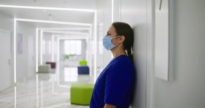 Female doctor in a blue coat takes off her mask and exhales after a difficult examination in a hospital corridor. Hard work of doctors in the hospital - Powered by Adobe