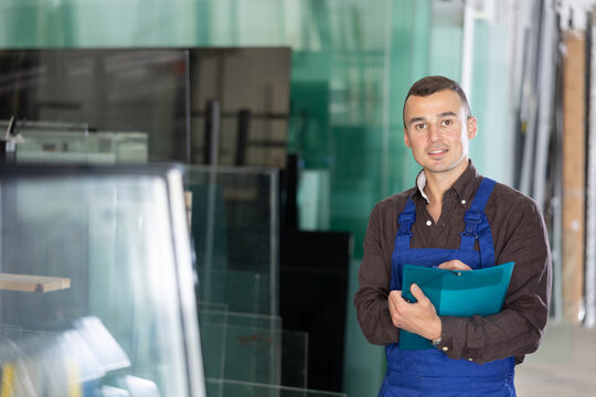 Positive male factory worker in blue overalls holding clipboard, standing confidently in workshop with glass panels in background..