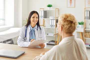 Fototapeta premium Smiling female doctor in white coat with stethoscope sitting at desk, holding clipboard, talking to elderly patient, providing professional medical consultation in bright organized medical office.