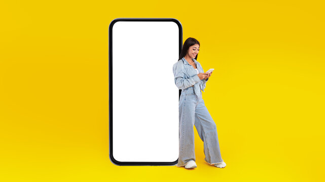 A young woman stands beside a large blank screen while texting on her smartphone. The bright yellow background creates an energetic and vibrant atmosphere perfect for digital content. - Powered by Adobe