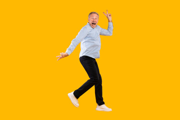 Excited mature man leaps high in a vibrant yellow orange studio, celebrating his success with a joyful expression and peace sign gesture, capturing the essence of triumph and happiness.