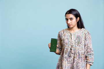 Obraz premium Happy indian woman presenting green screen on her smartphone, posing against the blue background. Joyous girl holding copy space device used for advertising brands in a commercial.