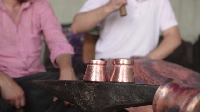 Traditional romani craftsman hammering a piece of copper on an anvil. Close-up on two handmade copper glasses with a blurred artisan at work