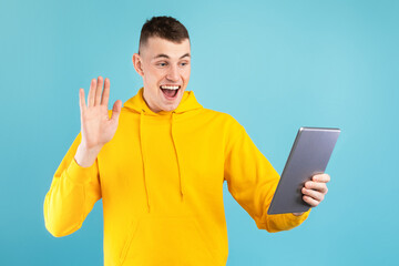 Excited young man in a yellow hoodie is smiling and waving at someone while holding a tablet. The background is a vibrant blue, creating a cheerful atmosphere.