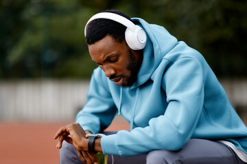 Focused black male athlete wearing headphones checks his time or pulse on a smartwatch after an intense workout on an outdoor running track.