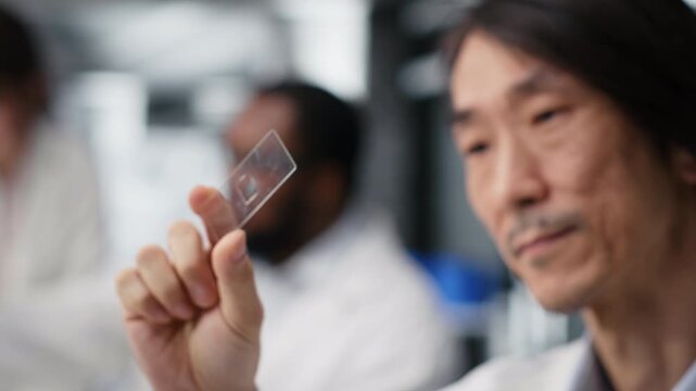 Senior microscopist examining microscope glass slide in laboratory to view cells. Asian research facility employee checks sample slide before imaging process using optical device, camera B