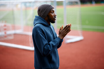 Focused black male athlete wearing a hoodie and a beanie rubs his hands together to warm up or concentrate before starting a workout on an outdoor sports field © djile