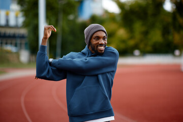 Smiling black male athlete happily stretches his triceps or shoulders as part of his energetic warm-up routine on an outdoor running track.