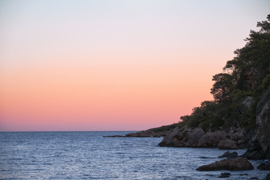 Tranquil sunset over Alacasu Paradise Bay (Cennet Koyu) near Kemer, Turkey. Warm pastel sky above the calm Mediterranean Sea and rocky coastline.