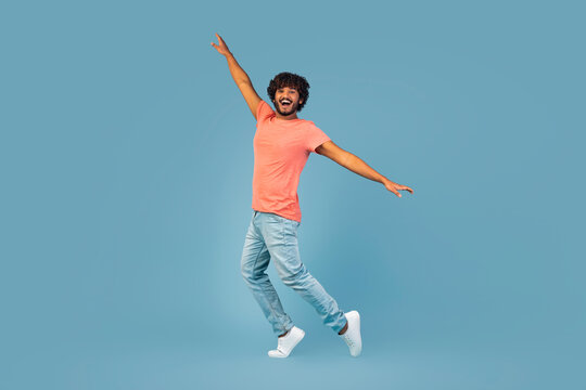 A cheerful young Hindu man in a casual outfit is dancing and jumping with joy against a blue studio background. He has a big smile and radiates positive energy with his movement.