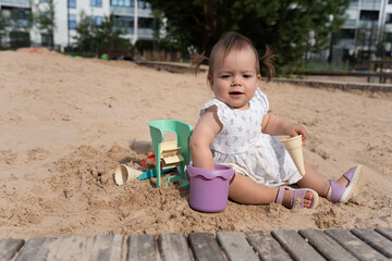 adorable toddler in a summer dress playing joyfully with colorful sand toys on a sunny day in a city playground surrounded by urban greenery and apartment buildings, relaxed, expressive, explore