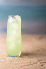 chilled green beverage in a tall glass with ice, set against a serene ocean-inspired backdrop, evoking a refreshing summer beach vibe on sandy textured surface, contrast, minimalism, idyllic, breeze