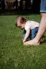 curious toddler bent over investigates grass with adult nearby, barefoot in sunlit park setting, emphasizing a playful and carefree outdoor atmosphere, happy, life, warmth, calm, horizontal, man