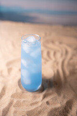 tall glass of refreshing blue beverage with ice cubes sits on sandy beach under soft light, evoking a cool and serene beachside atmosphere with gentle waves in the background, warmth, lifestyle