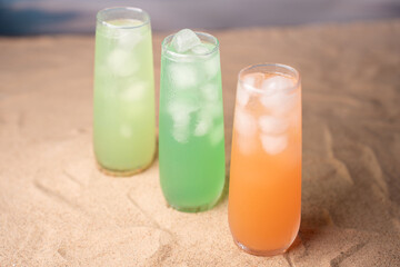 three refreshing tall glasses of vibrant colorful drinks with ice cubes on sand, capturing a tropical and refreshing beach atmosphere with soft natural lighting, easygoing, relax, chill, delight