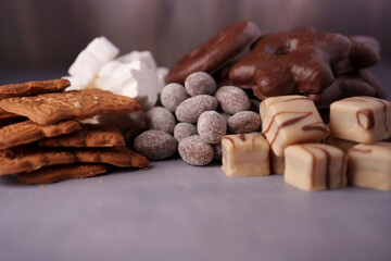 assortment of diverse confectionery items including biscuits, marshmallows, chocolate-covered treats, sugar-dusted nuts, and white chocolate cubes on a soft gray surface, baked, arrangement