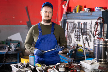 Positive male auto mechanic in blue overalls holding metal tool in car workshop