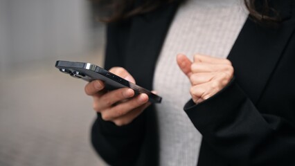 Caucasian female standing outdoors around grey modern buildings, working on her phone, smiling, touching her long dark hair. Close up.