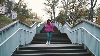 Smiling pretty woman in stylish jacket talking on the smartphone while walking down the stairs in the park. Seasonal style, autumn season concept. © Vlad