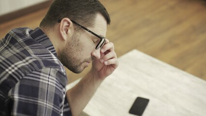 Tired young man taking off glasses, sitting in room at home. Phone on the table