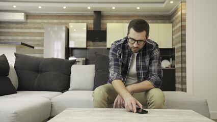Obraz premium Upset young man in glasses using smartphone while sitting on the sofa at home