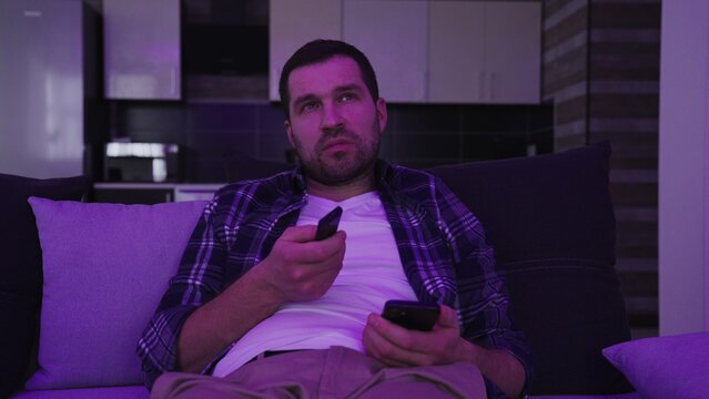Bored young man sitting on couch and watching tv in living room at night