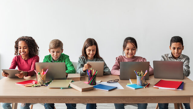 Diverse school children are seated at desks, using laptops and tablets for their studies. They appear happy and focused in a bright, modern classroom environment.