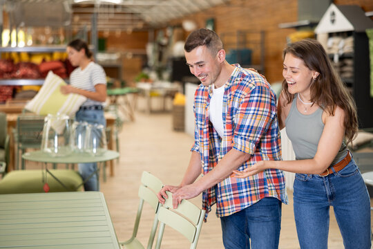 Cheerful young couple thoughtfully evaluating patio furniture options in garden store, choosing comfortable and functional chairs - Powered by Adobe