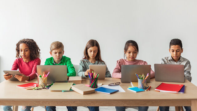 Five multiethnic school children are engaged in an online class while sitting at a desk. They actively use laptops and tablets for learning in a contemporary classroom environment.