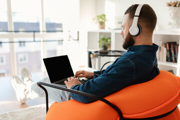 A young man sits in an orange chair, focused on his laptop. He wears headphones and enjoys a...