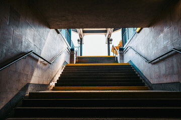 View from the bottom of a stairway leading up to street level in an urban subway or underpass. Sunlight brightens the exit framed by concrete walls, metal railings, and a hint of sky above