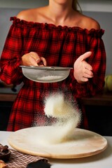 Woman in red plaid dress sifting flour over wooden surface in a cozy kitchen, showcasing baking preparation and culinary creativity with vibrant textures