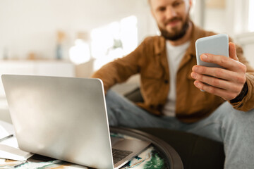 A man sits cross-legged in a comfortable workspace holding a smartphone. A laptop is placed on a table in front of him, suggesting he is multitasking or engaged in work.