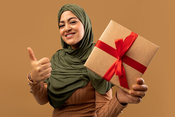 Muslim woman holding gift box and showing thumb up, wearing hijab and smiling, posing over brown studio background. Positive arab lady holding birthday or Christmas present