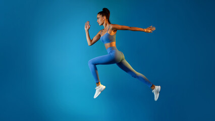 Obraz premium A determined young sportswoman is captured in mid-air, leaping with confidence. She is dressed in a fitness outfit and appears serious as she poses against a vibrant blue background.