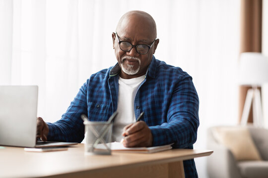 A mature African American man sits at a desk in a modern office, intently taking notes while using his laptop. He is focused on his work, wearing glasses and a plaid shirt.