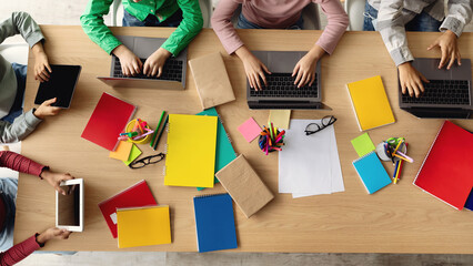 Brightly colored notebooks, supplies, and laptops are seen as school kids work together indoors. Students engage in learning and browsing the internet as they complete assignments.