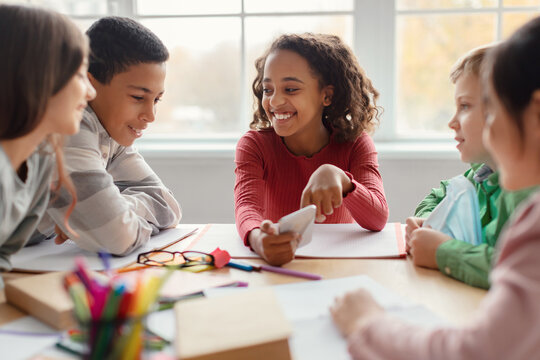 Multiethnic school children are having fun during their break in a classroom. They gather around a smartphone as an African American schoolgirl shows a new educational app to her classmates.