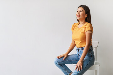 Asian lady smiles while showing her shoulder with a band aid after getting a COVID vaccine. She looks confidently into the empty space against a clean white background.