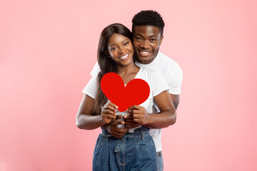 In a joyful moment, a young black couple embraces while holding a red paper heart. They smile at the camera, showcasing their love in a pink studio setting, perfect for Valentine's Day.