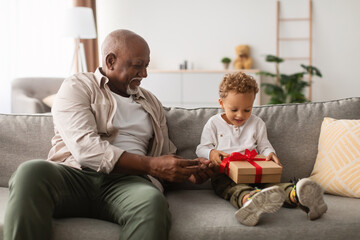 A joyful grandfather gives a wrapped birthday gift to his little grandson sitting on a couch at...