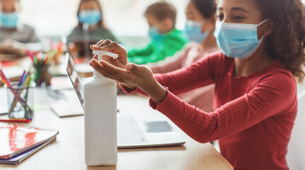 In a modern classroom, a Black schoolgirl is applying hand sanitizer to disinfect her hands while sitting at a table with classmates.