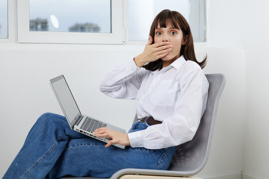 A surprised young woman sits on a modern chair with a laptop on her lap, hand covering her mouth, expressing shock and disbelief during online work or unexpected news.