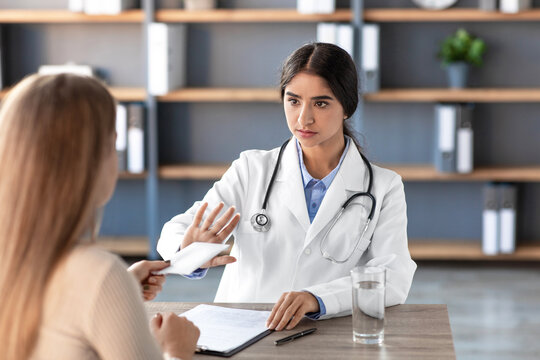 In a modern clinic office, a strict Indian woman doctor in a white coat firmly refuses a payment from her client. The scene highlights the importance of integrity in health care.