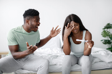 In a bright bedroom, a couple sits on a bed, visibly expressing strong emotions. The man appears excited while the woman looks distressed, holding a pregnancy test.