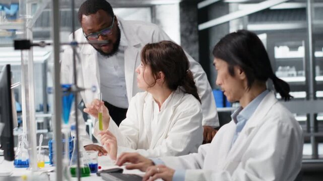 Multiethnic research facility team using colorful chemical solutions in glassware. Diverse teamworking analytical chemists in laboratory looking at reagent vial used for experiment, camera B