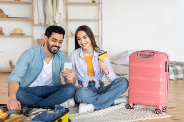 A couple is excitedly discussing their travel plans while surrounded by luggage and travel items. They are using a phone and a credit card to make bookings in a cozy, well-lit space.