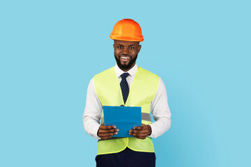 Professions Concept. Handsome Black Engineer In Hard Hat And Vest Holding Clipboard And Smiling At Camera, Portrait Of Young African American Construction Manager Posing On Blue Studio Background